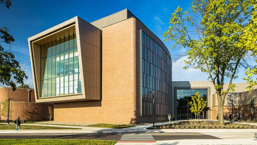 A brick and glass university building with a projecting glass corner, seen from the street on a clear day.