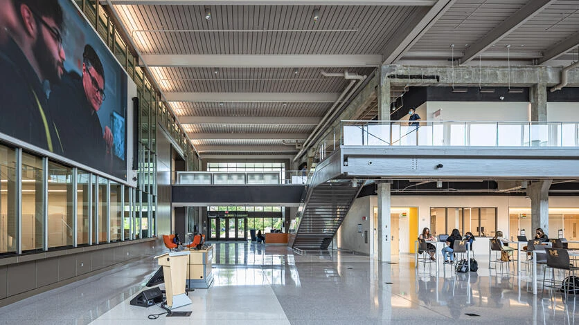 A two-story atrium inside the building, with a staircase, a walkway above, and students working at tables.